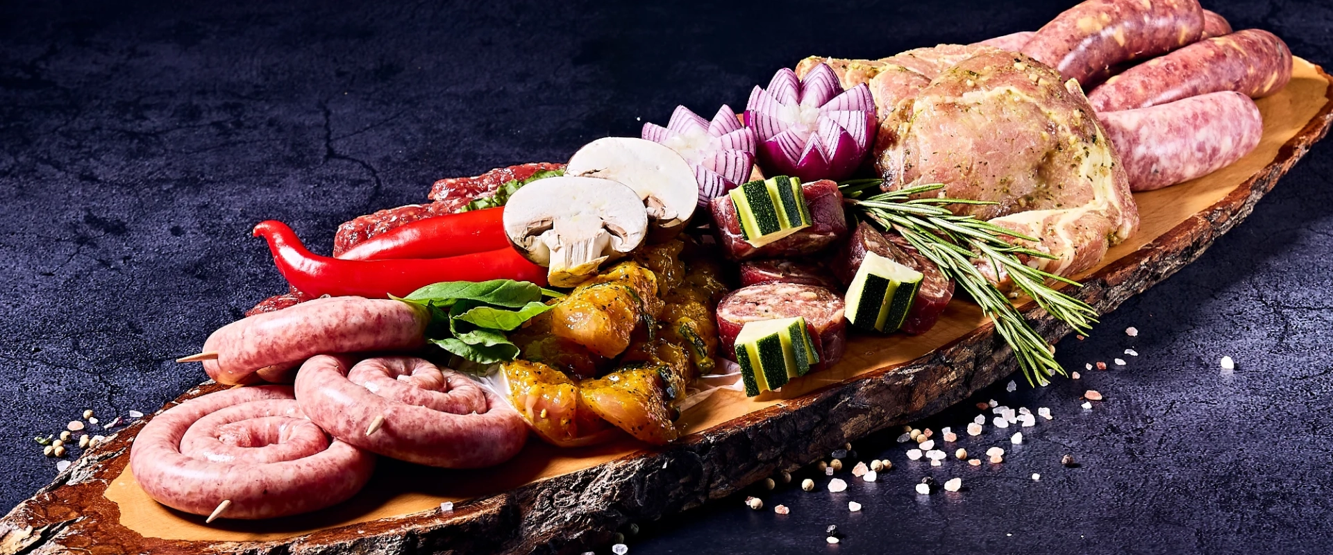 Metzgerei Thumm Filderstadt – Hintergrund – Grillplatte Holzplatte mit einer Auswahl an rohem Grillfleisch, Bratwürsten, marinierten Fleischspießen, Zucchini, Paprika, Champignons, Zwiebeln und frischen Kräutern, dekorativ angerichtet auf dunklem Untergrund.