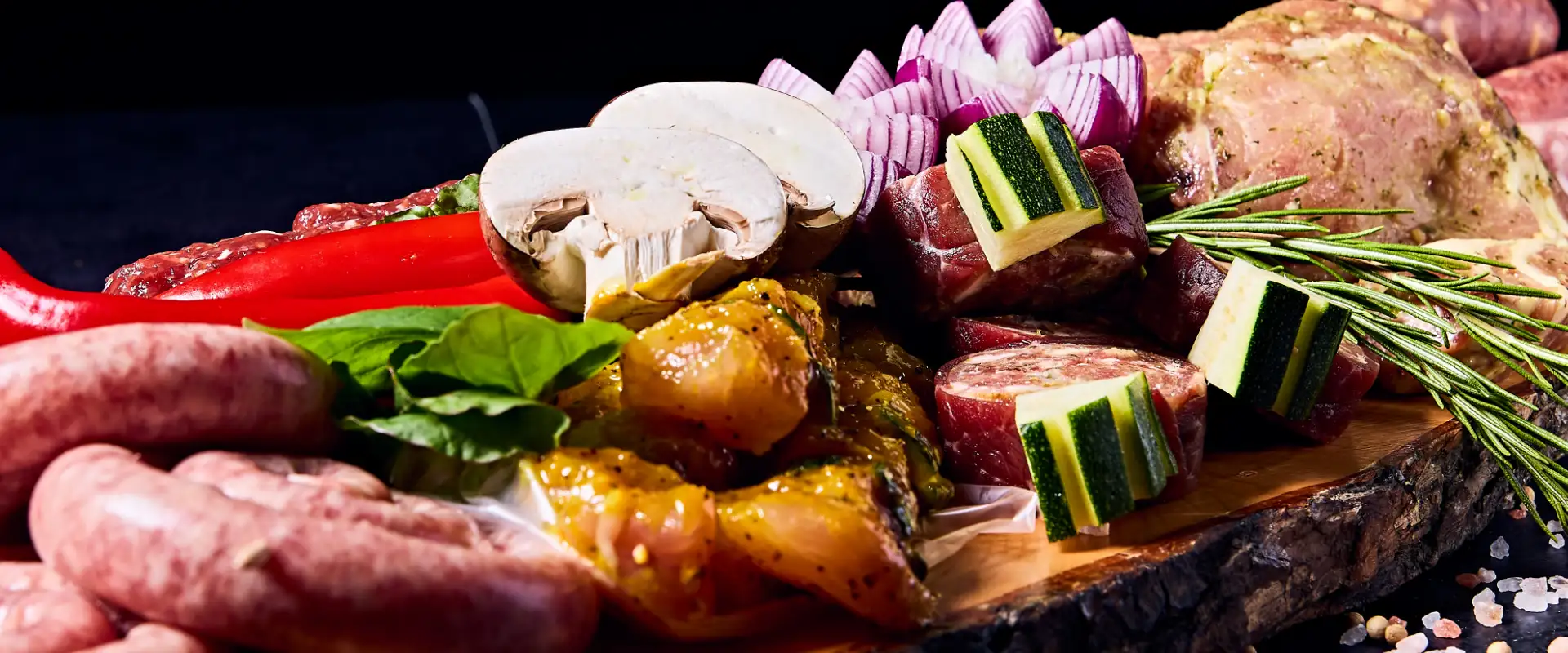 Metzgerei Thumm Filderstadt – Hintergrund – Partyservice Nahaufnahme einer rustikalen Holzplatte mit rohen Grillzutaten wie Bratwurst, mariniertem Fleisch, Gemüsespießen mit Zucchini, Champignons, roten Zwiebeln, Paprika und frischen Kräutern auf dunklem Untergrund.